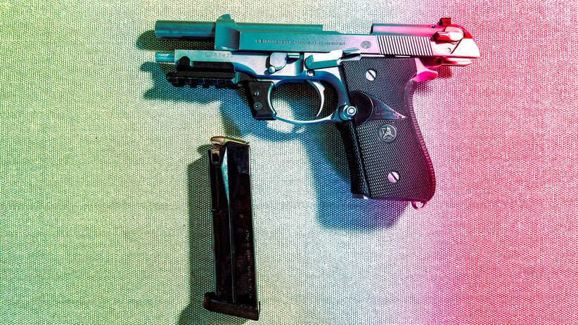 A silver pistol with the magazine ejected, lying below the trigger. The mechanism is pulled back, revealing the barrel and chamber. A soft rainbow gradient lights the image from left to right.