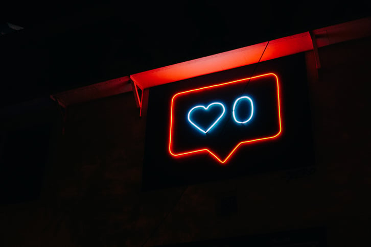 A red-and-blue lit neon sign in the darkness: a blue heart next to a zero, outlined by a red speech box. The neon sign is hanging from a red rafter.